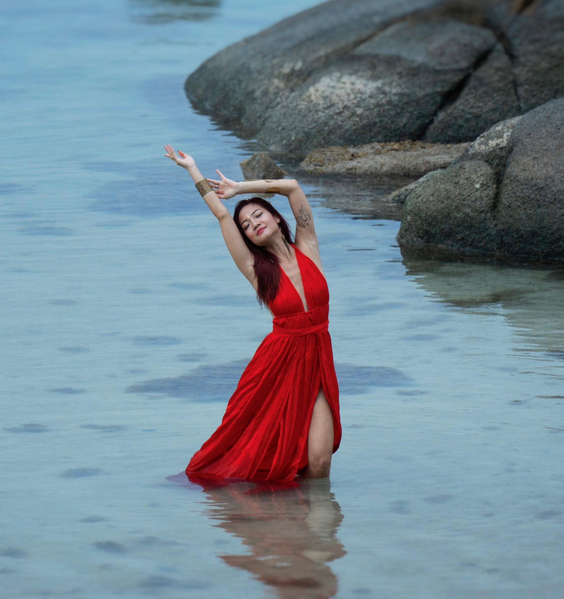 Janel Aurelia in a flowing red dress, kneeling in shallow ocean water