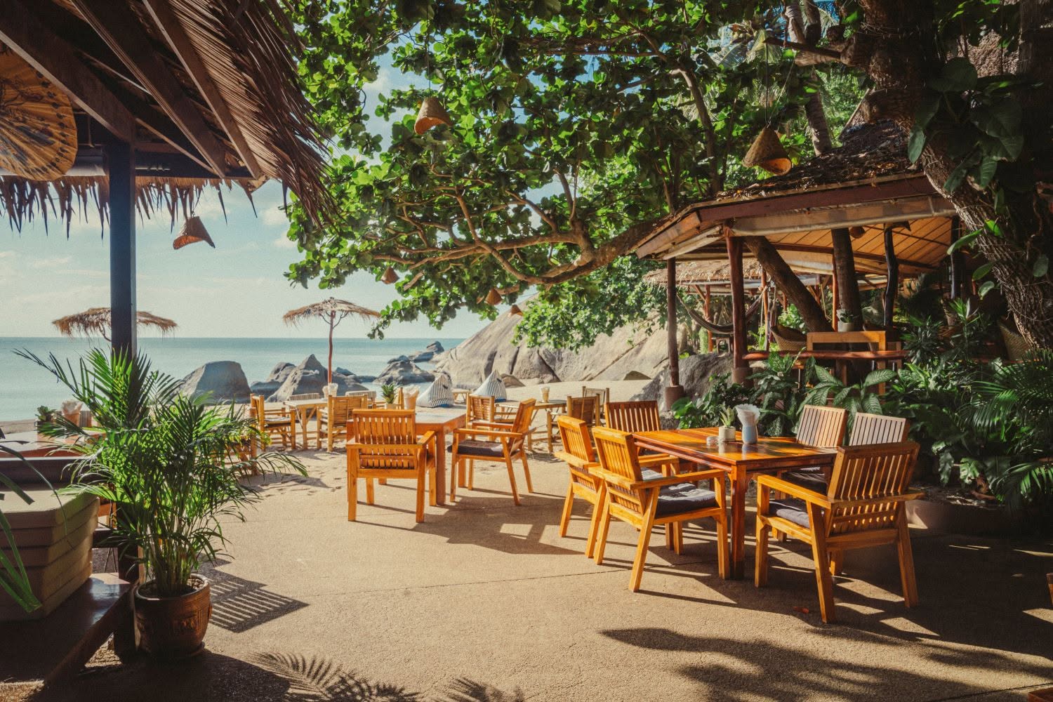 Open-air beachfront dining with wooden tables under trees and thatched roofs